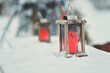 Rustic wooden lanterns on tables outside, red candles inside the lanterns. Tables are covered in snow and lanterns are sitting on top of the snow surface, one lantern in focus. Snowy winter day.