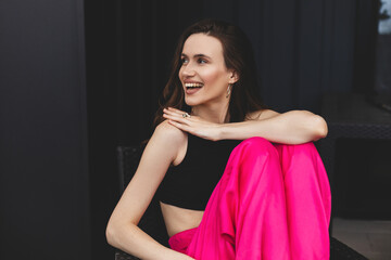 Poster - Attractive brunette woman sitting at home outdoors, wear pink pants, black top circle earrings. Girl joy of free time, look happy, smiling, look bright. Shot of happy hippie lady isolated black wall.