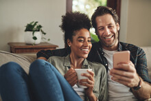 Look At Whats Trending Online Today. A Happy Young Couple Using A Cellphone Together While Relaxing On A Couch At Home.