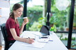 © ND STOCK - Smiling businesswoman holding coffee cup and working on laptop in office