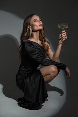 Sticker - Fashionable beautiful brunette woman with diamond earrings in a stylish elegant evening black dress with a glass of champagne sits and poses in a studio with light. Chic Lady celebrates birthday event