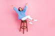© deagreez - Full length photo of gorgeous pretty girl blue sweater white pants sitting on bar stool raising arms up isolated on pink color background