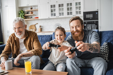 Wall Mural - KYIV, UKRAINE - APRIL 19, 2023: Smiling gay parent playing video game with daughter near partner and drinks at home