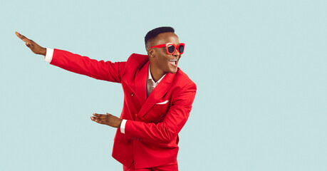Happy young man in stylish party outfit dancing and having fun in studio. Cheerful excited African American man in red suit and sunglasses dancing isolated on light blue background. Fashion concept