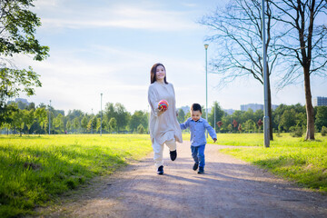 Asian mother and child play with ball, run across the field, on the grass. Kazakh family together in holiday. Happy woman and boy in summer pastime. daylight saving time