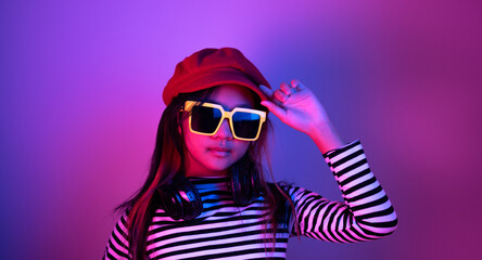 Portrait of a young Asian girl with yellow sunglasses and ear loops around her neck and a red hat with blue and red neon lights.