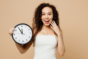 Happy fun satisfied young woman bride wearing wedding dress posing hold in hands clock cover mouth isolated on plain pastel light beige background studio portrait. Ceremony celebration party concept.