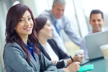 She adds brains and beauty to the organization. a young Asian businesswoman sitting at a conference table with her colleagues in the background.