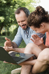 Wall Mural - happy father pointing at laptop next to teenage boy while sitting together on green lawn.