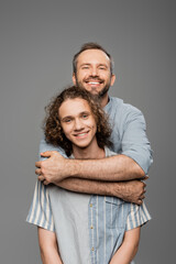 Wall Mural - cheerful man hugging curly teenager son and looking at camera isolated on grey.