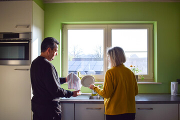 Mature couple washing dishes together in kitchen. Sharing time together. Rinsing and drying plates. People in the morning smiling. Solar panels in the background. Morning routine together.