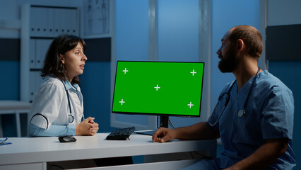 Wall Mural - Medic and assistant looking at computer monitor with green screen chroma key isolated display while discussing patient disease expertise. Medical team working late at night in hospital office