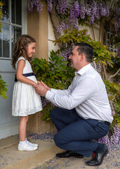 Father and daughter. A little girl in a white dress is standing, dad kneeled in front of her. Look into each other's eyes. Father's love and care for his daughter Flower background, wisteria.