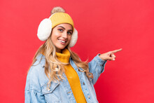 Young Pretty Uruguayan Woman Wearing Winter Muffs Isolated On Red Background Background Pointing Finger To The Side