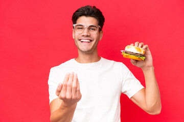 Wall Mural - Young Caucasian man holding a burger isolated on red background inviting to come with hand. Happy that you came