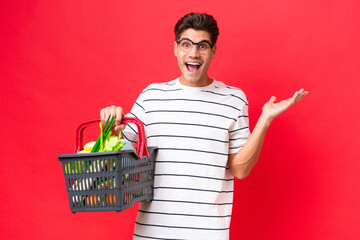 Young caucasian man man holding a shopping basket full of food isolated on red background with shocked facial expression