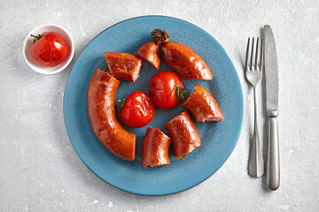 Wall Mural - Fried pork sausage cut into pieces in a blue plate with tomatoes and cutlery on a light gray stone background