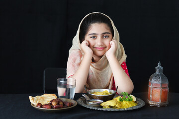 Wall Mural - Adorable happy Muslim girl with beautiful eyes wearing hijab and traditional costume resting chin in hands at kitchen table, portrait of kid eating Islamic halal food meal on dark black background.