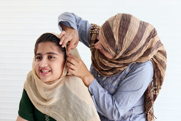 Wall Mural - Muslim mother dressing up her daughter girl with traditional hijab scarf, adorable happy smiling kid with beautiful eyes smiling at mom, happy lovely Islamic family on white background.