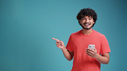 Young latin or arabian man using smartphone standing very happy pointing with hand and finger to the side, in coral t-shirt on blue studio background