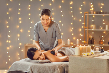 Wall Mural - Woman getting hot stones massage at massage salon or spa room with lights and candles. Relaxed young woman lying on spa bed while professional masseuse puts hot stones on her back