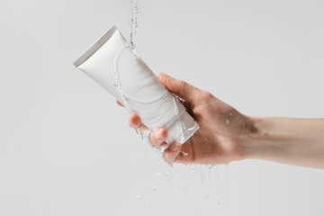 Female hand holding white mockup tube of moisturizer under water on white isolated background. Concept of face care cosmetics, anti-wrinkle cream concept.