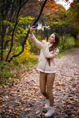 Wall Mural - Woman taking selfie in autumn park. Young attractive lady using smartphone.