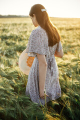 Wall Mural - Beautiful woman in floral dress walking n barley field in sunset light. Stylish female holding straw hat and relaxing in evening summer countryside. Atmospheric moment, rustic slow life