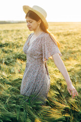 Wall Mural - Beautiful woman in floral dress walking n barley field in sunset light. Stylish female in straw hat and relaxing in evening summer countryside. Atmospheric moment, rustic slow life