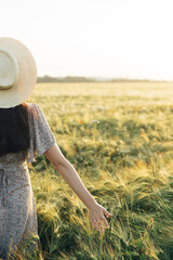 Wall Mural - Beautiful woman in floral dress walking in barley field in sunset light. Atmospheric moment, rustic slow life. Stylish female holding straw hat and enjoying evening summer countryside
