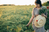 Fototapeta Sypialnia - Beautiful woman with wildflowers and straw hat walking in barley field in sunset light. Stylish female relaxing in evening summer countryside and gathering flowers. Atmospheric tranquil moment