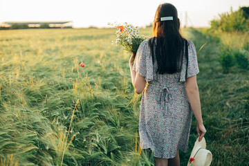 Wall Mural - Beautiful woman with wildflowers and straw hat walking in barley field in sunset light. Stylish female relaxing in evening summer countryside and gathering flowers. Atmospheric tranquil moment