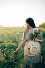 Wall Mural - Beautiful woman with straw hat and wildflowers enjoying sunset in barley field. Atmospheric tranquil moment, slow life. Stylish female gathering flowers and walking in evening summer countryside