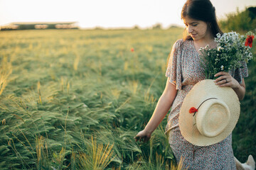 beautiful woman with wildflowers and straw hat walking in barley field in sunset light. stylish fema