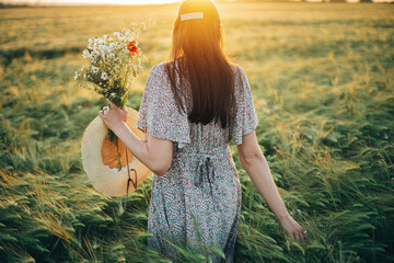 Wall Mural - Beautiful woman with wildflowers bouquet standing in barley field in sunset light. Stylish female relaxing in evening summer countryside and gathering flowers. Atmospheric tranquil moment