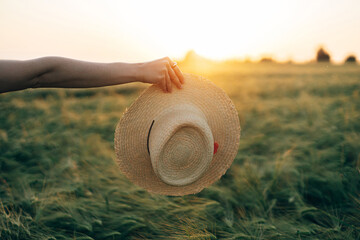 Wall Mural - Straw hat in woman hand in sunset light on background of barley field. Stylish female enjoying evening summer countryside. Atmospheric tranquil moment, rustic slow life