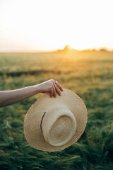 Wall Mural - Woman holding straw hat in sunset light on background of barley field. Stylish female relaxing in evening summer countryside. Atmospheric tranquil moment, rustic slow life