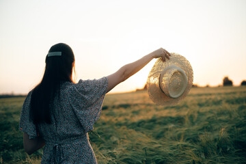 Wall Mural - Woman holding straw hat in sunset light on background of barley field. Stylish female relaxing in evening summer countryside. Atmospheric tranquil moment, rustic slow life