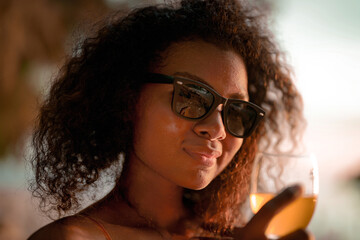 Wall Mural - African woman drinking cocktail juice when sitting on bench beach at tropical beach. Young traveler wearing yellow bikini and chilling out the beauty of the Nature. Wanderlust and travel concept.