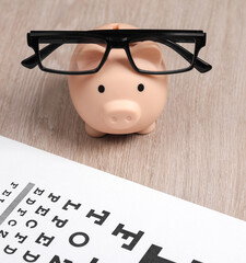 Wall Mural - Piggy bank in Eyeglasses with eye test chart on table. Vision examination and correction. Top view