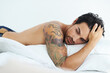 © Cameron Mcdonald/peopleimages.com - Thinking, relax and shirtless with a sexy man lying on a bed in studio on a white background. Tattoo, idea and topless with a handsome young male model posing in a bedroom for sensuality or desire
