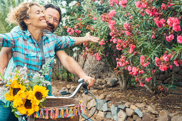 Wall Mural - Exited couple in outdoor leisure activity riding a bike together with red flowers in background. Woman outstretching arms and man laughing. People enjoying green transport together and happy lifestyle