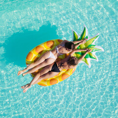 Wall Mural - Top view of two girls relaxing on inflatable big pineapple in the pool. Aerial view of ladies relaxing on the floating mattress.