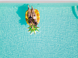 Wall Mural - Top view of two girls relaxing on inflatable big pineapple in the pool. Aerial view of ladies relaxing on the floating mattress.