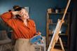 © Andrii Lysenko - Brazilian woman artist works in concentration. Side view portrait of a creative lady paints a picture on canvas at an easel in an art studio at home. tired stands and wipes sweat from her face