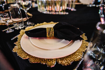 Serving, setting table. Plate decorated linen black napkin and silverware cutlery, glasses. Side view. Closeup. Wedding set up, dinner table reception. Birthday, baptism, event. Luxury golden decor.