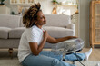 Overheating in homes. Happy young African American woman sits in front of electric fan on floor in living room, enjoying blowing cool air, overheated female cooling down at home after being in heat
