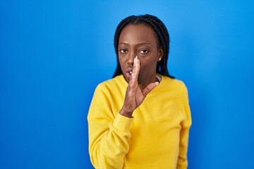 Wall Mural - Beautiful black woman standing over blue background hand on mouth telling secret rumor, whispering malicious talk conversation