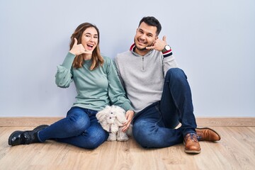 Canvas Print - Young hispanic couple sitting on the floor with dog smiling doing phone gesture with hand and fingers like talking on the telephone. communicating concepts.