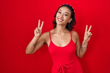 Wall Mural - Young hispanic woman standing over red background smiling looking to the camera showing fingers doing victory sign. number two.